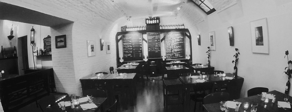 casaluigi Schwarz-Weiß-Foto eines gemütlichen, gedämpft beleuchteten Restaurants mit ordentlich angeordneten Tischen, Kerzen auf jedem Tisch, weißen Backsteinwänden, gerahmten Bildern und einer großen Speisekarte auf der Rückseite.