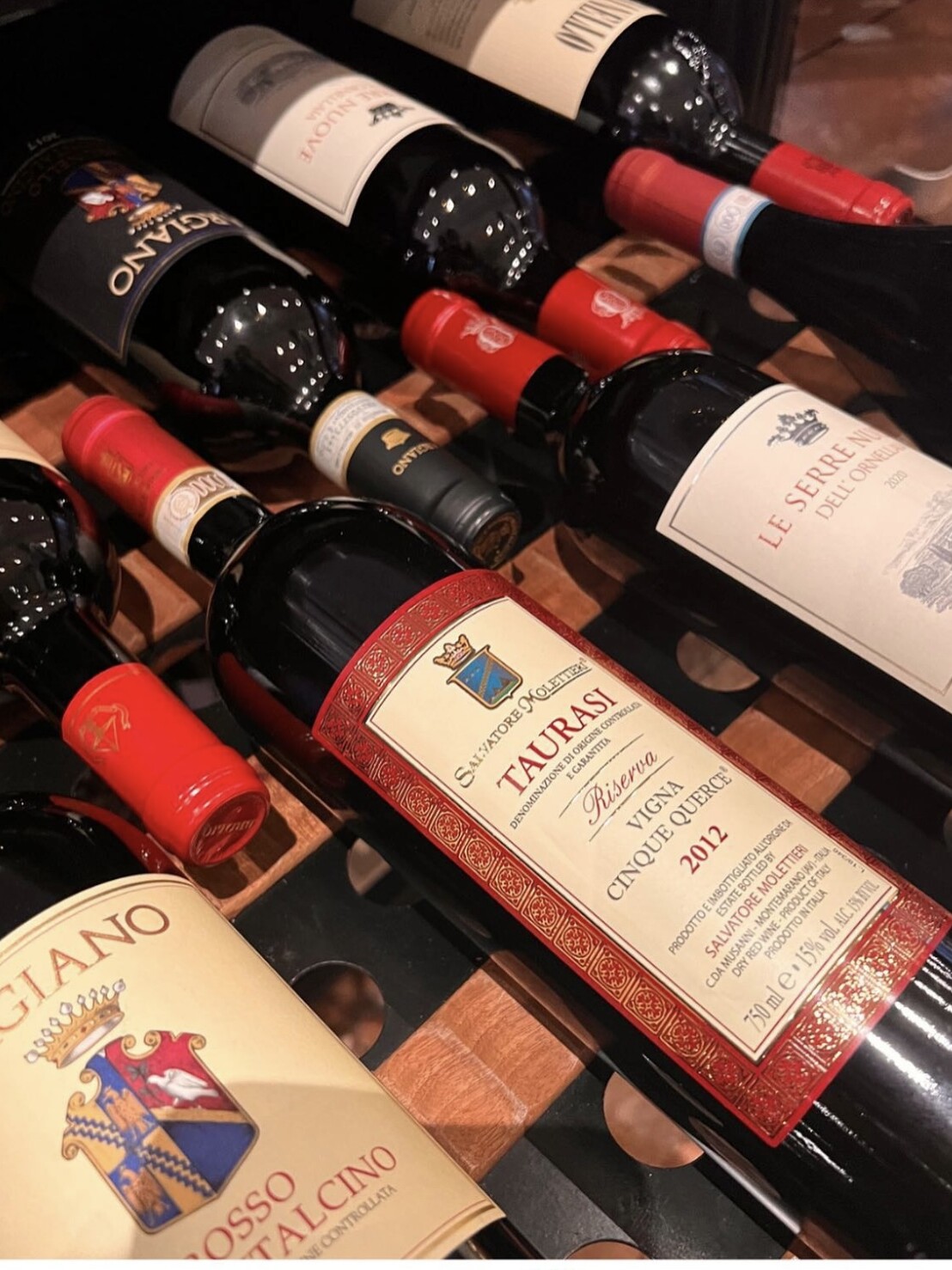 casaluigi Eine Nahaufnahme mehrerer Rotweinflaschen auf einem Weinregal mit Etiketten verschiedener italienischer Weingüter, darunter Taurasi, Rosso di Montalcino und Le Serre Nuove dell'Ornellaia.