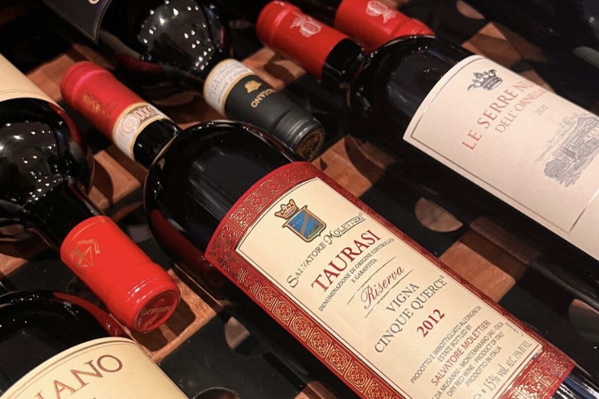 casaluigi Eine Nahaufnahme mehrerer Rotweinflaschen auf einem Weinregal mit Etiketten verschiedener italienischer Weingüter, darunter Taurasi, Rosso di Montalcino und Le Serre Nuove dell'Ornellaia.