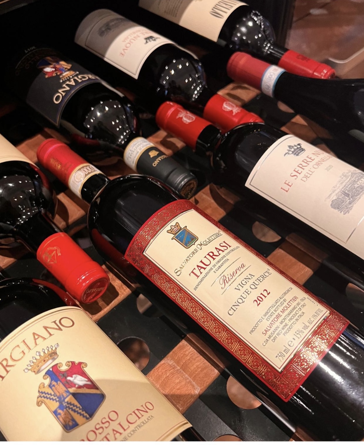casaluigi Eine Nahaufnahme mehrerer Rotweinflaschen auf einem Weinregal mit Etiketten verschiedener italienischer Weing&uuml;ter, darunter Taurasi, Rosso di Montalcino und Le Serre Nuove dell'Ornellaia.
